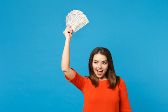 Beautiful brunette young woman wearing red orange dress holding fan of cash money in dollar banknotes isolated over blue wall background, studio portrait. People lifestyle concept. Mock up copy space.