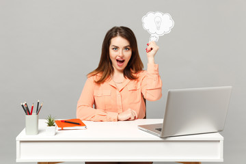 Young surprised woman in pastel clothes holding say cloud with lightbulb sit work at desk with pc laptop isolated on gray background. Achievement business career lifestyle concept. Mock up copy space.