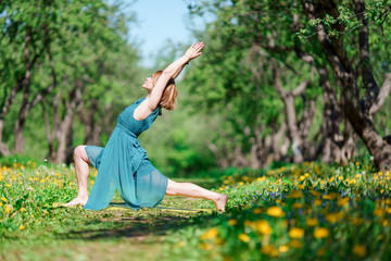 Photo on side of young woman with raised arms in long green dress doing yoga in forest