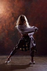 Fototapeta na wymiar Photo of young dancing blonde girl in ripped jeans and sneakers on dark brown background in studio