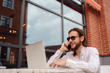 Photo of cheerful brunet man freelancer talking on phone while sitting at table with laptop near brick building on summer day