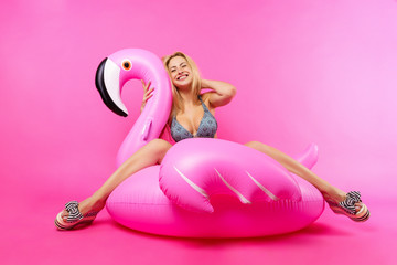 Image of smiling blonde woman in bathing suit and pink glasses sitting on inflatable flamingo on...