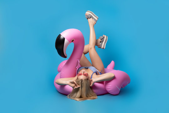Image of blonde in bathing suit and in pink glasses lying on back on pink inflatable flamingo on empty blue background
