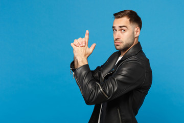 Handsome stylish young unshaven man in black leather jacket white t-shirt holding hands like gun, side view isolated on blue background studio portrait. People lifestyle concept. Mock up copy space