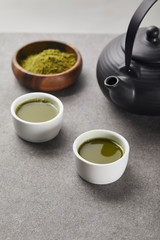 selective focus of green matcha powder in wooden bowl near cups with tea and black teapot