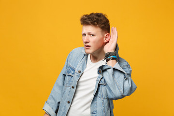 Portrait of curious young man in denim casual clothes try to hear you with hand near ear isolated...