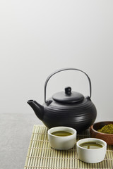 selective focus of green matcha powder in wooden bowl near black teapot and cups with tea on bamboo...