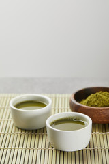 selective focus of green matcha powder in wooden bowl near white cups with tea on bamboo table mat