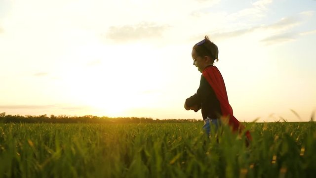 The camera follows the running child against the sunset. A charming laughing boy runs on green grass in a superhero costume.