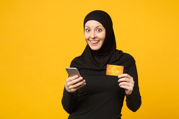 Young arabian muslim woman in hijab black clothes hold in hand mobile phone, credit card isolated on yellow wall background, studio portrait. People religious lifestyle concept. Mock up copy space.