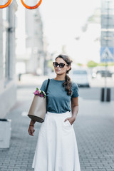 Obraz premium Fashionable young woman in sunglasses, a white skirt and a blue blouse walks city with a bag and flowers. European retro style.