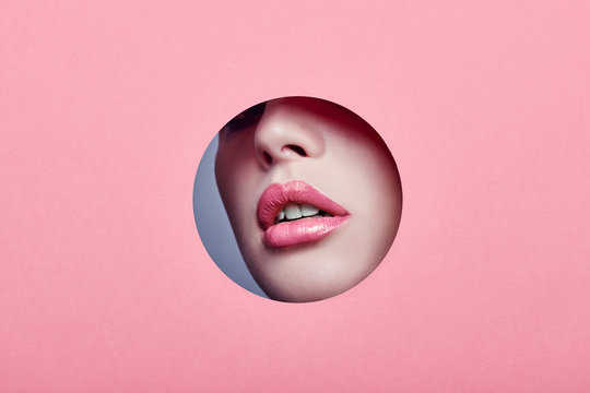 Advertising Beautiful plump lips bright pink color, woman looks in hole colored pink paper, beauty salon. Makeup, advertising face care, perfect lips, Fashion beauty makeup and cosmetics