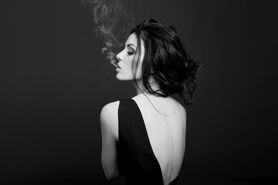 Fashion Beauty Makeup Cosmetics Brunette Woman Smokes On Dark Background In Black Dress, White Smoke From Her Mouth. Erotic Girls, Black And White