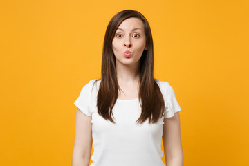 Obraz premium Portrait of pretty funny young woman in white casual clothes looking camera, blowing lips isolated on bright yellow orange wall background in studio. People lifestyle concept. Mock up copy space.