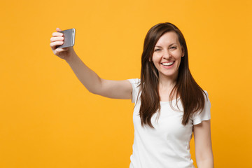 Portrait of laughing young woman in white casual clothes doing selfie shot on mobile phone isolated on bright yellow orange wall background in studio. People lifestyle concept. Mock up copy space.