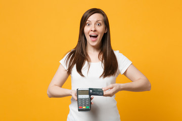 Surprised young woman holding wireless modern bank payment terminal to process and acquire credit card payments isolated on yellow orange wall background. People lifestyle concept. Mock up copy space.