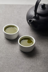 selective focus of black teapot and white cups with matcha tea on grey stone table