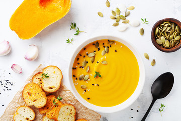 Autumn pumpkin cream soup in bowl served with seeds and crouton on white stone table top view.