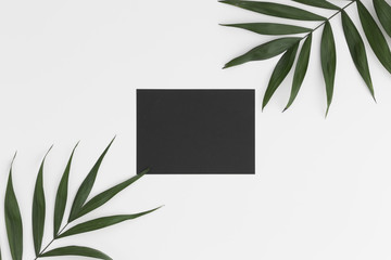 Top view of a black card mockup with palm leaf decoration on a white table.