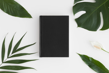 Top view of a black book mockup with tropical leaves decoration on a white table.