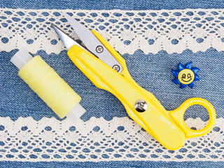 White lace, yellow thread and scissors, blue button on jeans background. Stitching still life