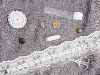Tailor's tools – white lace, threads, scissors, ribbon, buttons, pins, thimble, tape measure on a background of gray fabric. Stitching still life