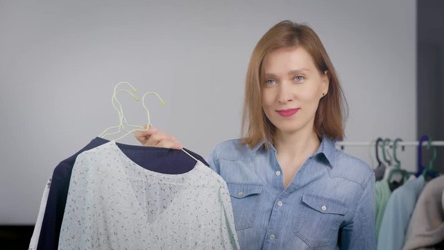 personal stylist woman is looking at camera and smiling, holding hangers with clothes and showing them