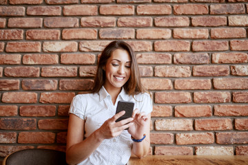 Beautiful smiling brunette sitting at coffee shop and using smart phone. In background brick wall.