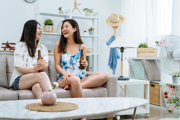 two happy female friends drinking beer talking at home in comfort temperature living room. young girls laughing gossip with electric fan turning on in apartment. beautiful ladies in modern house