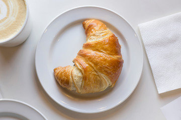 Fresh croissant on the white plate, cappuccino, napkin on the table. Top view, copy space