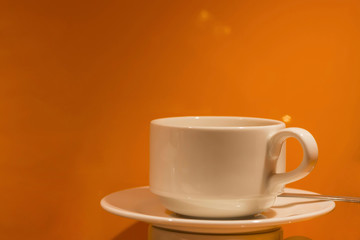 White cup and plate, spoon on dark orange background, highlights. Autumn, tea concept, close-up, copy space, toned