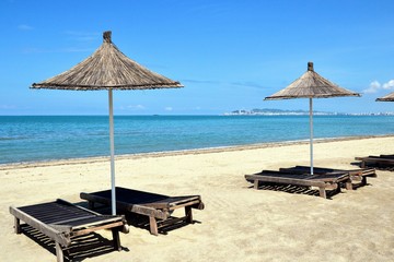 Umbrella and chaise lounges on the sandy beach. Wooden beach chairs, beds and straw umbrella on a tropical beach. Deck chairs and umbrella at the Albanian beach, Golem. Durres city in a distance