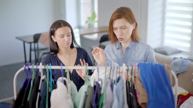 one woman is putting dress on hanger in room and her female friend are cutting threads with funny face