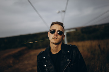 Young sexy man in leather jacket and sunglasses standing outdoor. Dark key photo