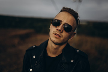 Young sexy man in leather jacket and sunglasses standing outdoor. Dark key photo