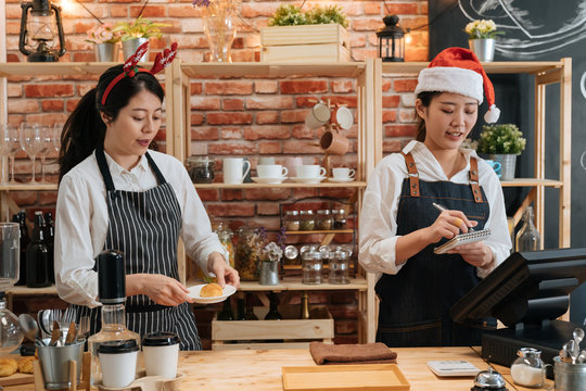 two asian female bartenders dressed in santa hat and working in bar counter in christmas holidays. woman barista prepare croissant while coworker waitress taking note on customer order in xmas.