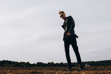 Young sexy man in leather jacket and sunglasses standing outdoor. Silhouette against grey sky