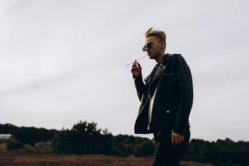 Young sexy man in leather jacket and sunglasses standing outdoor. Silhouette against grey sky