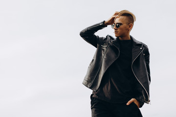 Young sexy man in leather jacket and sunglasses standing outdoor. Silhouette against grey sky