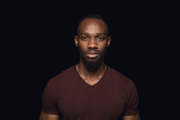 Close up portrait of young african man isolated on black studio background. Photoshot of real emotions of male model. Standing and looks serious. Facial expression, human nature and emotions concept.