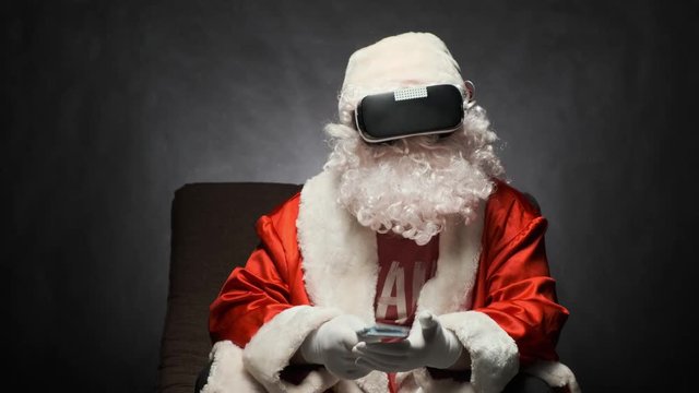 Satisfied Santa Claus holds a bundle of money in his hand while sitting on a sofa wearing virtual reality glasses.