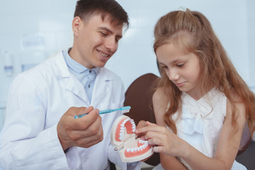 Young handsome male dentist showing his young patient how to brush teeth properly; using dental mold and a toothbrush. Cute young girl learning brushing teeth from professional dentist