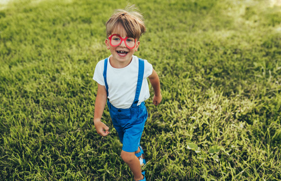 Outdoors image of happy little boy wearing blue shorts, white t-shirt and red paper glasses playing at nature background. Cheerful child running on the green grass in the park. Kid having fun.
