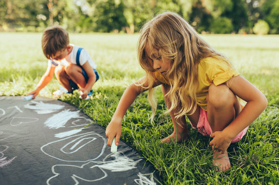 Children playing with colorful chalks. Happy little kids drawing with chalks in the park. Friends, boy and two girls having fun on sunlight outdoors. Childhood
