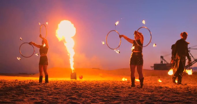 Fire show three women in their hands twist burning spears and fans in the sand with a man with two flamethrowers in slow motion.