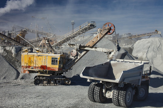 Heavy truck drives up to the excavator for gravel loading on the background of a rock stone crushing complex. Mining industry. Quarry mining equipment.