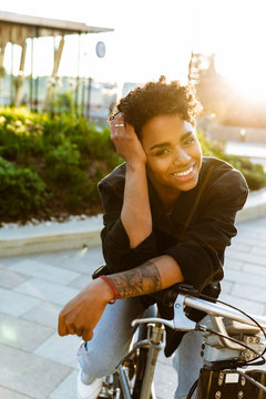 Photo of joyous african american woman smiling while sitting on bicycle in city park