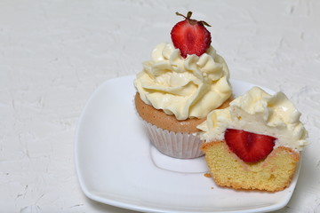 Cupcakes with strawberries and butter cream. One of them is cut, the filling is visible. On a white surface with decorative plaster.