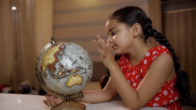 Thoughtful little Indian child preschooler looking and playing with a world globe in the study room. Indian stock footage of a female junior sitting in a casual outfit and revolving the globe