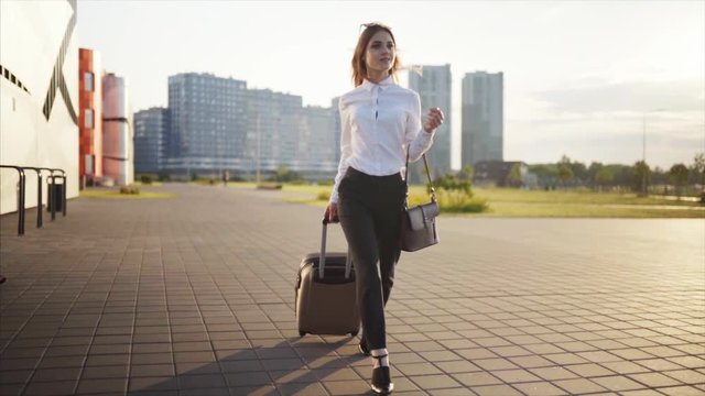 Slow motion. Business girl with hand-baggoes to the departure area, on the background developed city, business centers and skyscrapers. Dressed in a white shirt and black trousers. Style casual